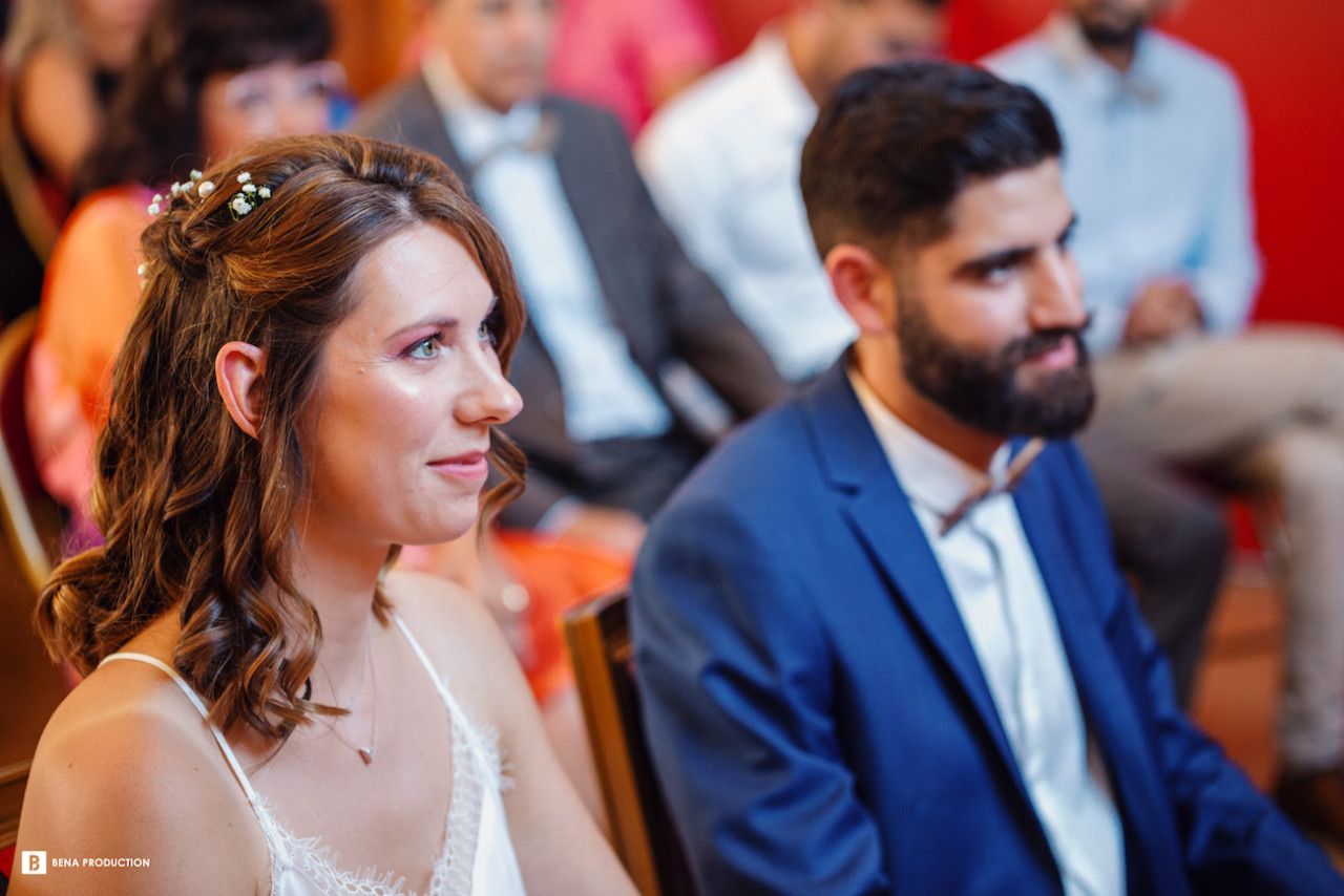 Nous découvrons Noémie et Benjamin assis à présent dans la salle des mariages de la mairie de Neuville-sur-Oise, très attentifs aux paroles de l'officier d'état civil qui célèbre leur mariage. Noémie, à la coiffure naturelle rehaussée d'un soupçon d'ornement floral? a laissé retomber ses boucles naturelles sur ses épaules. Un discret tour de cou orné d'une pierre orne son décolleté. Son bustier, délicatement ourlé et agrémenté de fines bretelles, met en valeur sa peau dorée. A ses côtés, Benjamin apparaît, radieux dans son costume bleu vif sur chemise immaculée, noeud papillon bien en place. Le jeune couple ne perd pas une miette des déclarations !