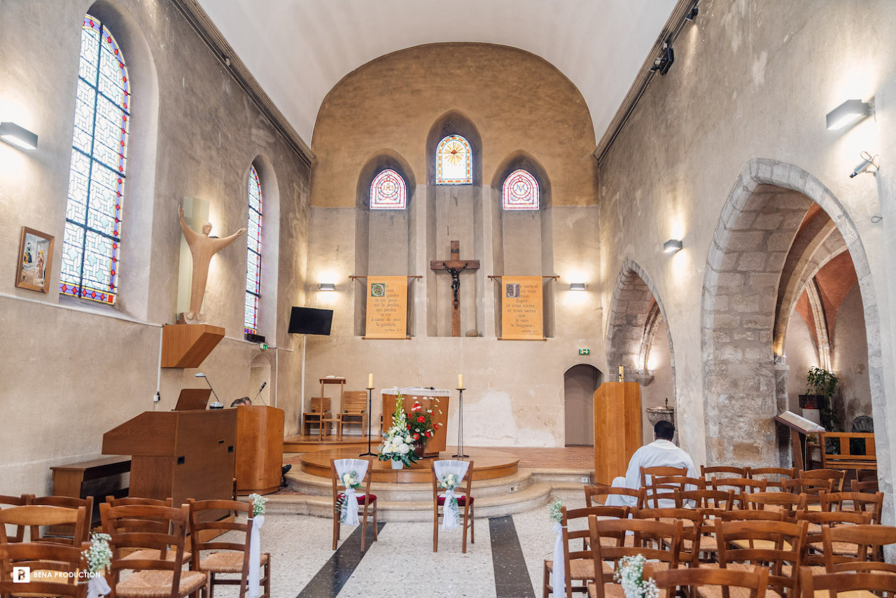 Nous découvrons l'inérieur de l'église, gaiement décorée e bouquets pour élébrer le mariage de Sylvie et Sylvain, largement éclairée par les vitraux et les nombreuses appliques sur les murs. Intérieur à la fois sobre et lumineux. On aperçoit le prêtre attendant les mariés au premier rang des chaises qui vous accueillir les familles et amis.